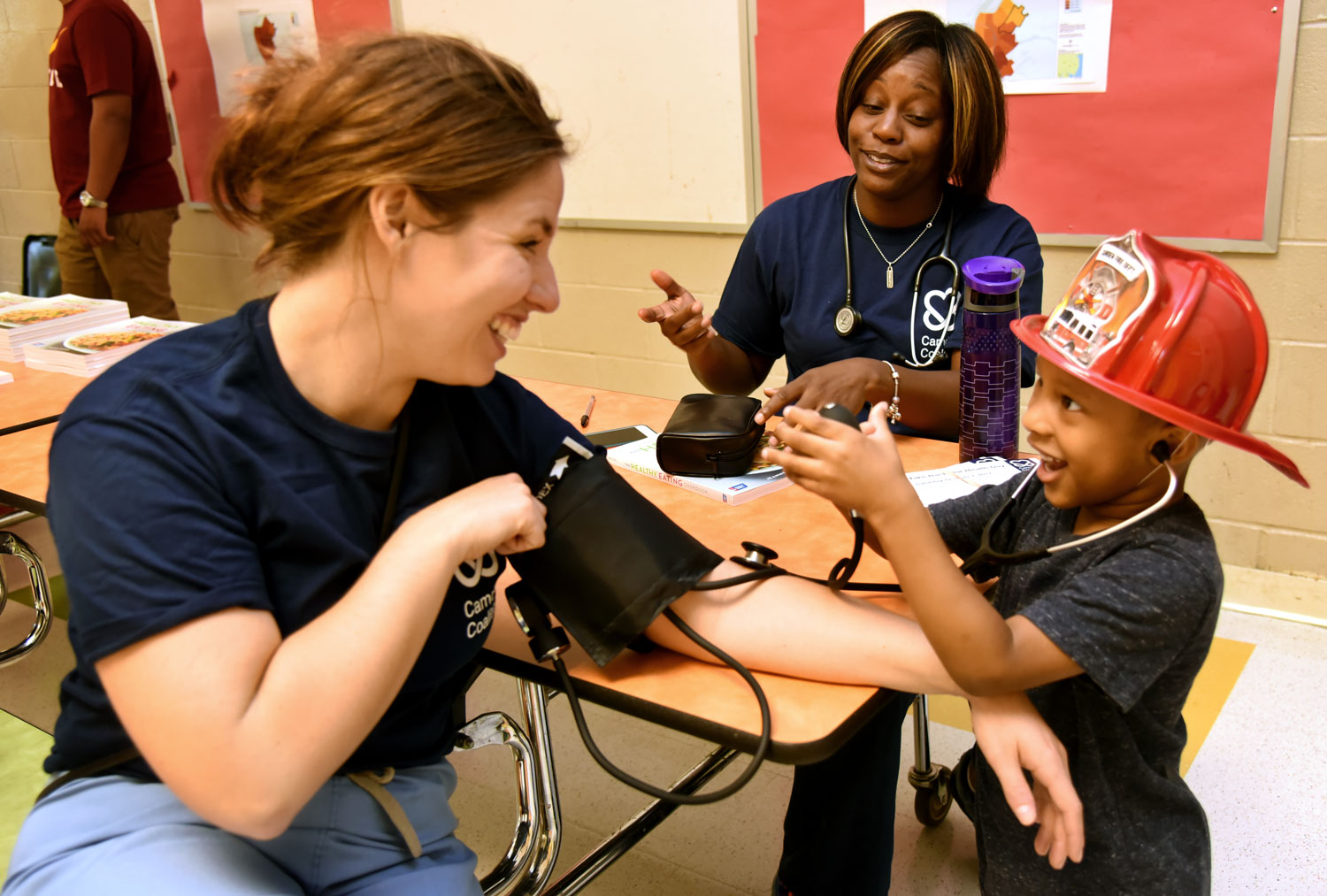 African American child in fireperson hat does blood pressure on two nurses