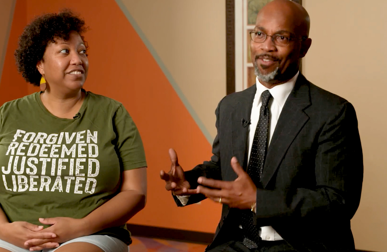 African American woman in "Forgiven Redeemed Justified Liberated" shirt looks at speaking African American man in suit.