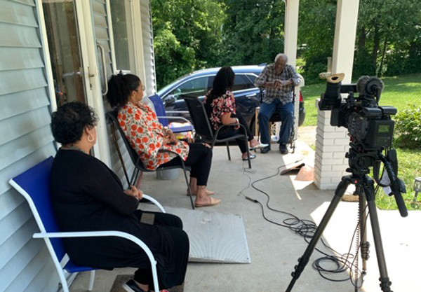 Photo on porch: elderly African American man is interviewed on camera by AA woman, while two other AA women watching