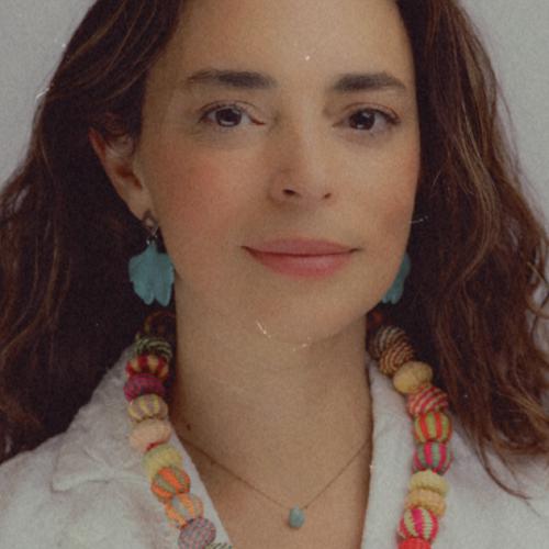 Uri Ramos headshot - fair-skinned woman with long brown hair wearing white shirt and colorfol necklace