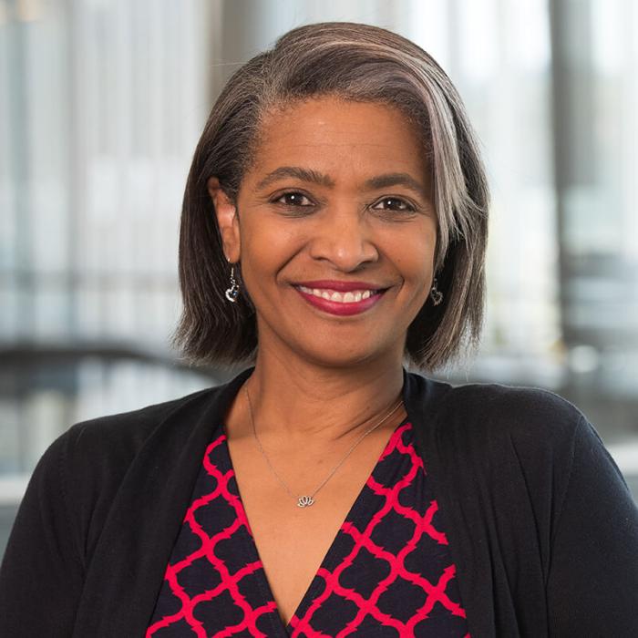 Headshot of Stephanie Yates - brown-skinned black woman with shoulder-length black and gray hair, a checkered blue and red shirt, and black blazer